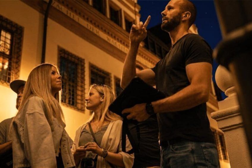 A group of people standing outdoors at night, with a building featuring classical architecture in the background. 3