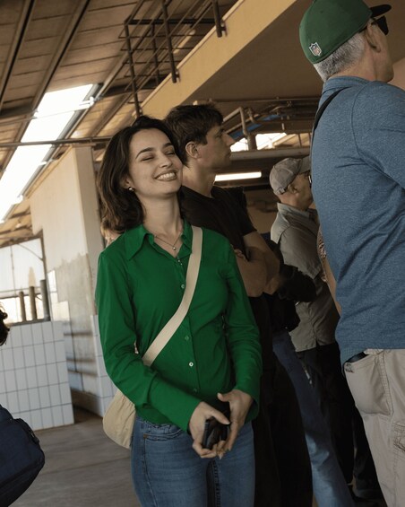 A woman in a green shirt and jeans standing in a covered outdoor area with a metal roof and tiled walls. 2