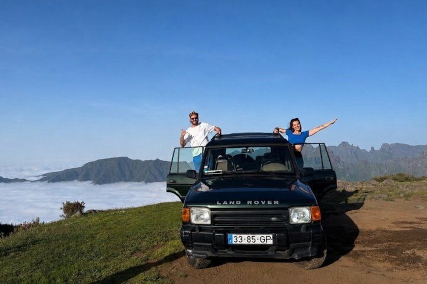 A black Land Rover with two people standing in the open door, a coastal landscape with mountains in the background. 1