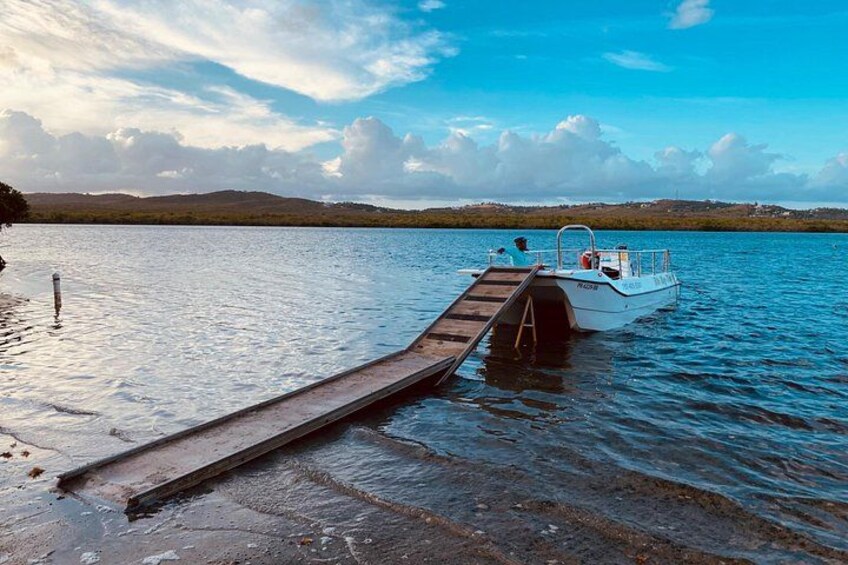 Un barco amarrado en un muelle que se extiende hacia aguas tranquilas, bajo un cielo despejado y colinas lejanas. 2