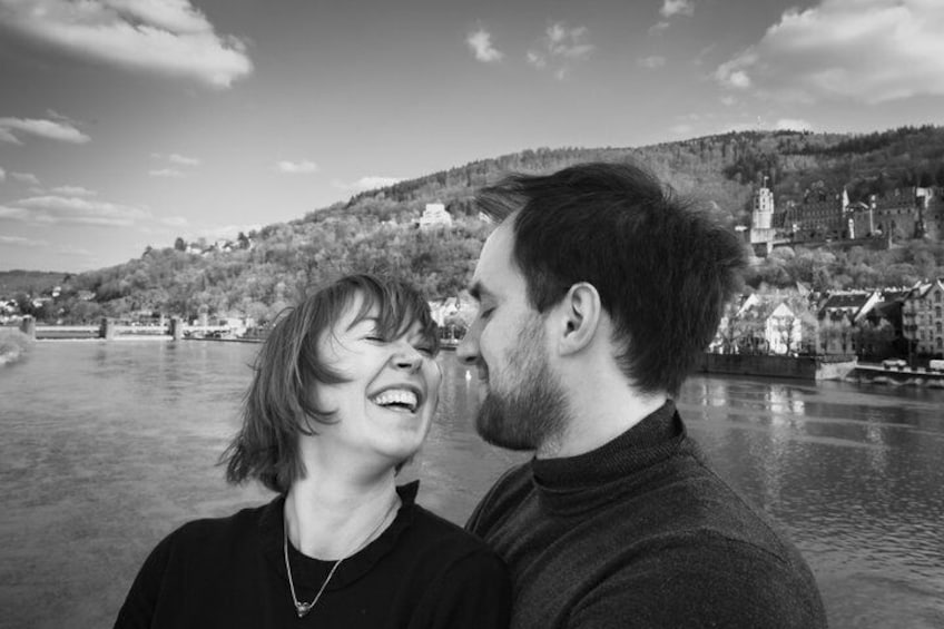 Un couple se tient au bord d’une rivière, avec une ville en arrière-plan. 1