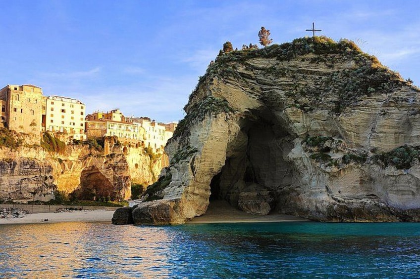 A coastal cliff with a cave entrance, a cross on top, and buildings in the background. 1