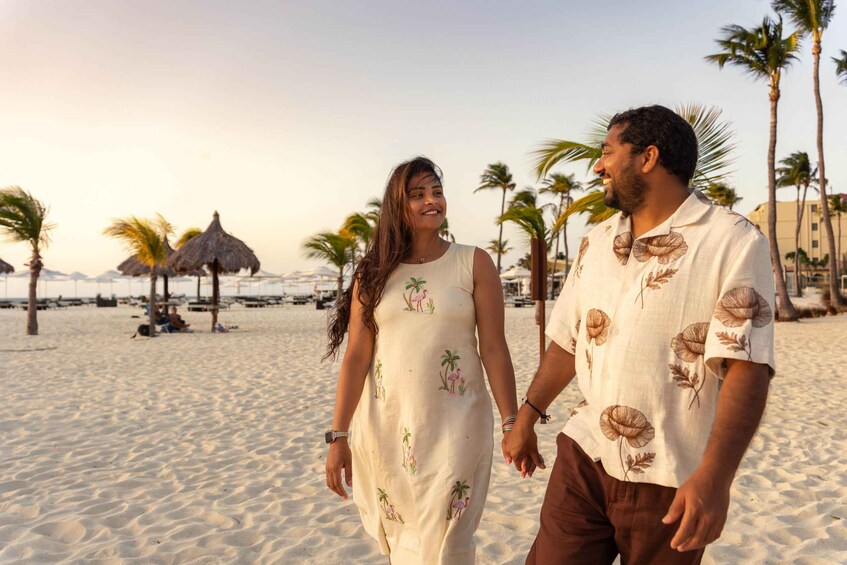A couple walking on a sandy beach with palm trees and thatched umbrellas in the background. 4