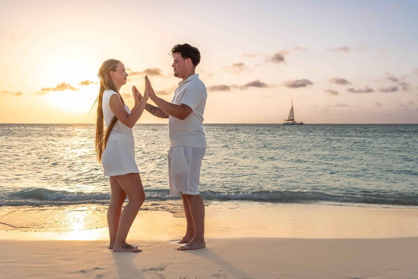 Two people standing on a beach at sunset with a sailboat in the background. 1