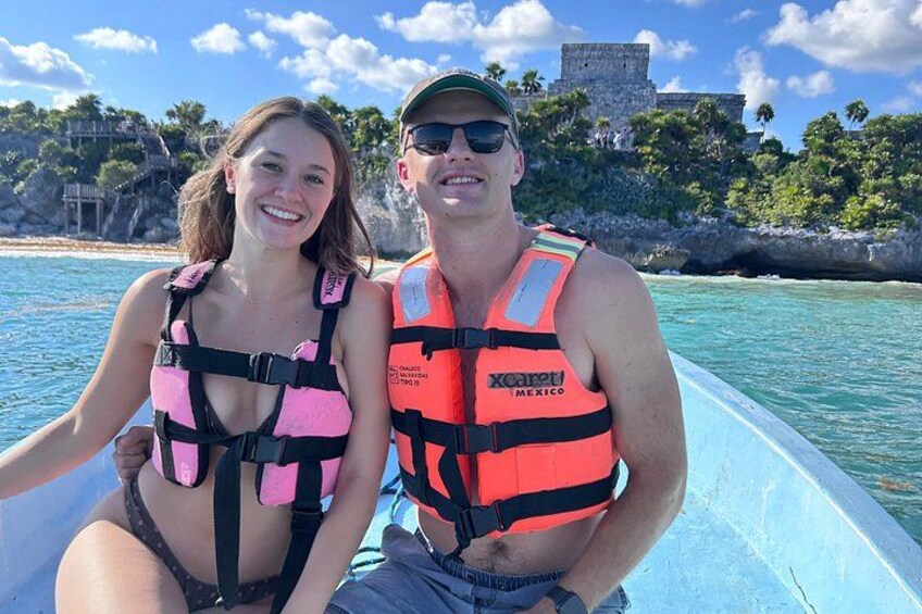 Two people on a boat wearing life jackets, with a coastal landscape and a historical building in the background. 3