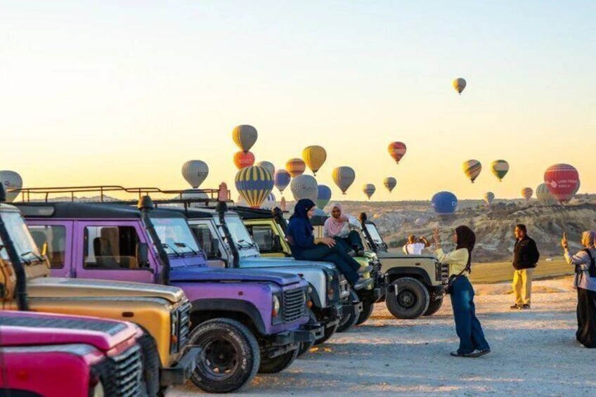 A group of people on safari vehicles, hot air balloons in the background, and a desert landscape. 1