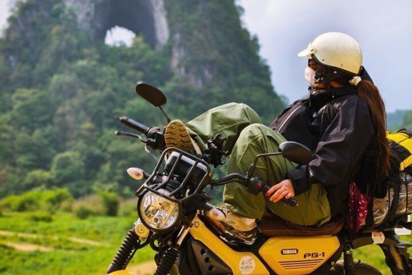 A person on a yellow motorcycle with a green bag, in a natural landscape with greenery and a rock formation in the background. 1
