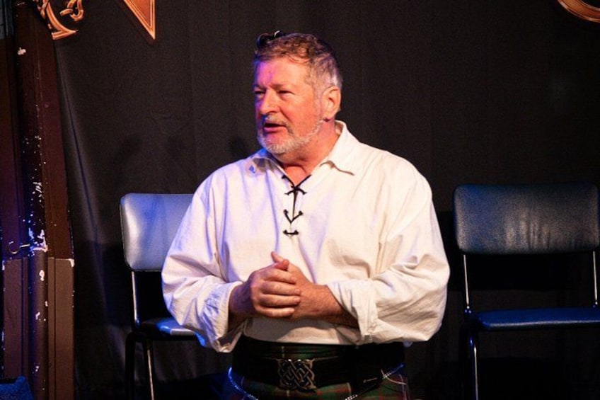 A man seated on a stage with a microphone, wearing a white shirt and a black belt. 2