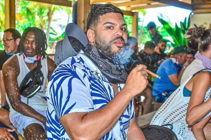 A person wearing a blue and white patterned shirt, holding a cigar, in a social setting with other people. 5