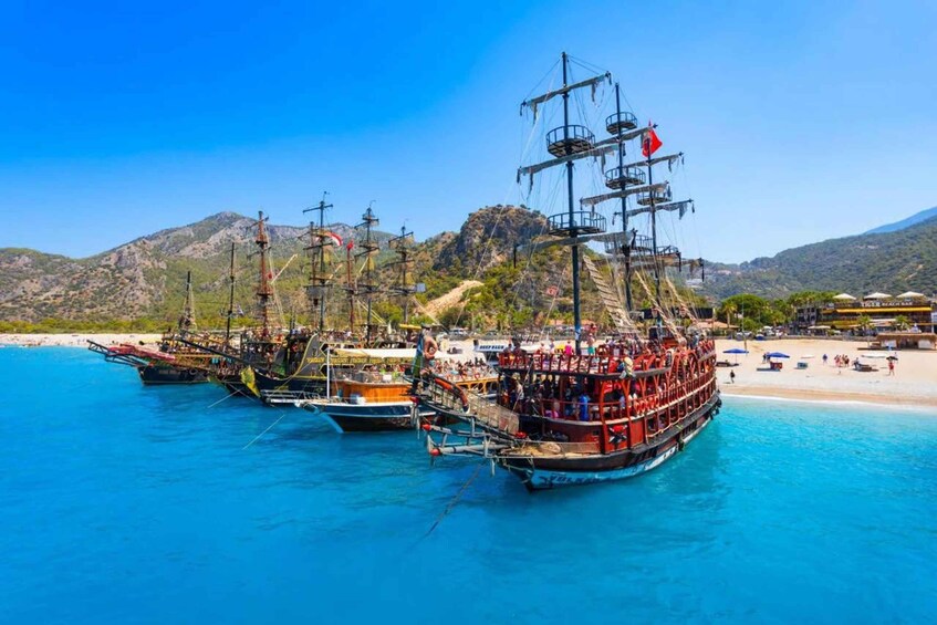 A fleet of sailing ships docked at a beach with clear blue waters and mountainous terrain in the background. 4