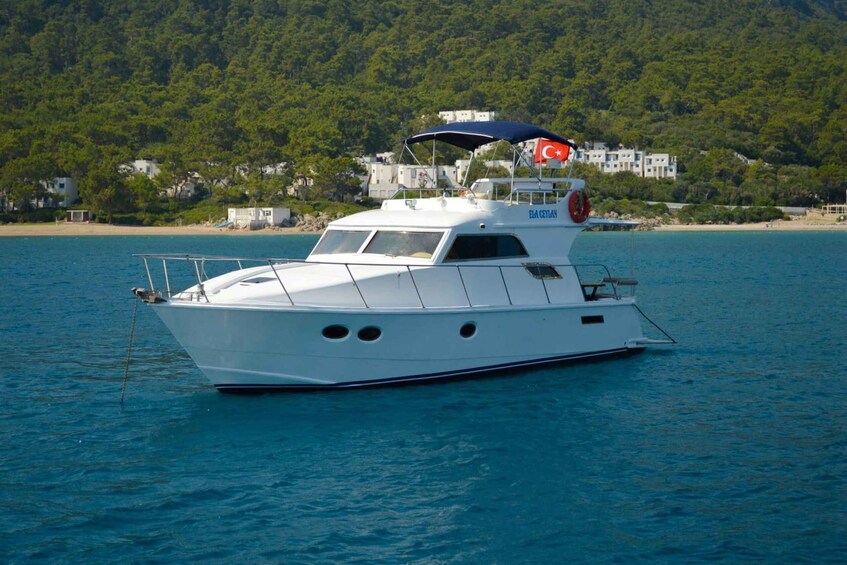 A white yacht with a Turkish flag on a calm sea, docked near a beach with houses and trees in the background. 5