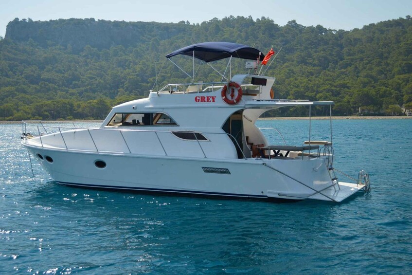 A white yacht named 'GREY' with a canopy, life preserver, and a forested shoreline in the background. 1