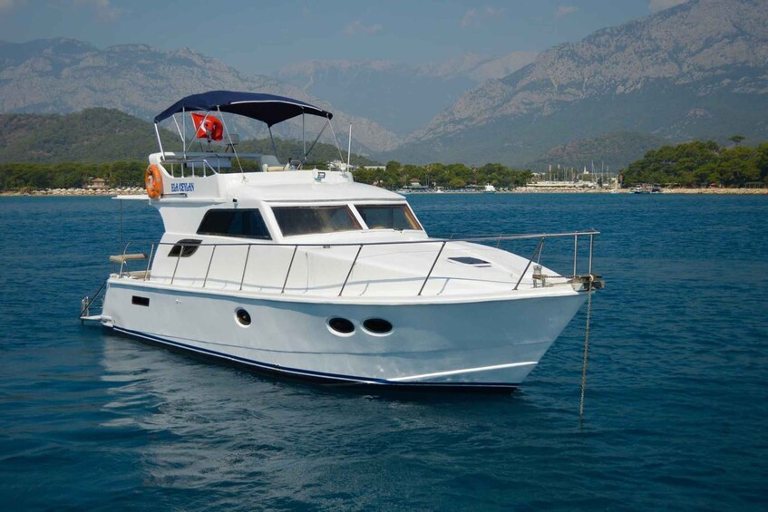 A white boat named 'GACELIAN' with a blue canopy and an orange life preserver on calm water with mountains in the background. 3