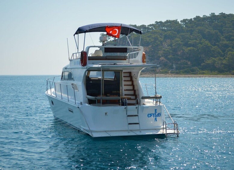 A white boat with a Turkish flag on the bow, sailing on calm waters with a forested coastline in the background. 2