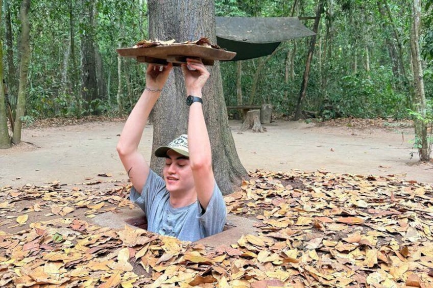 A person sitting on the ground surrounded by dry leaves, holding a tray above their head. 3