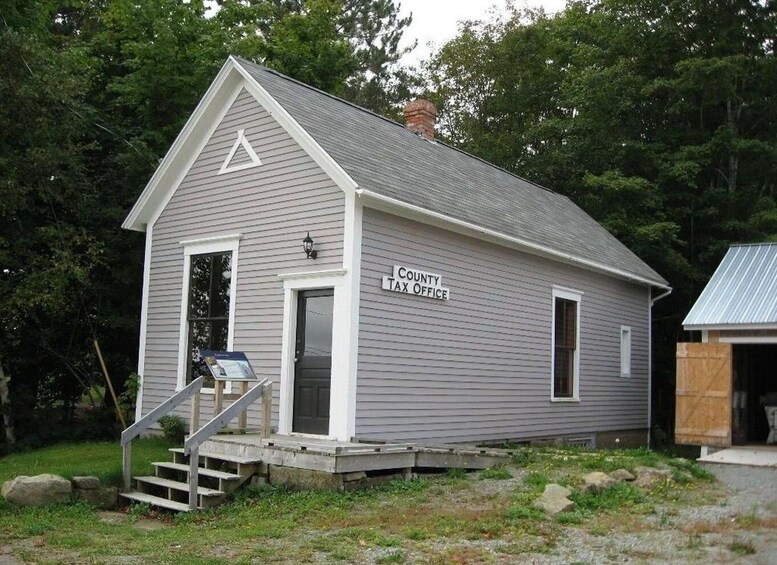 A small, single-story building with a gabled roof, labeled as a County Tax Office, surrounded by trees and a grassy area. 4