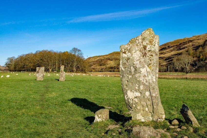 A stone circle with standing stones in a grassy field, surrounded by hills and trees. 2