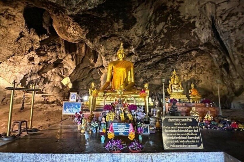 A cave with a golden Buddha statue, smaller statues, and offerings. 4