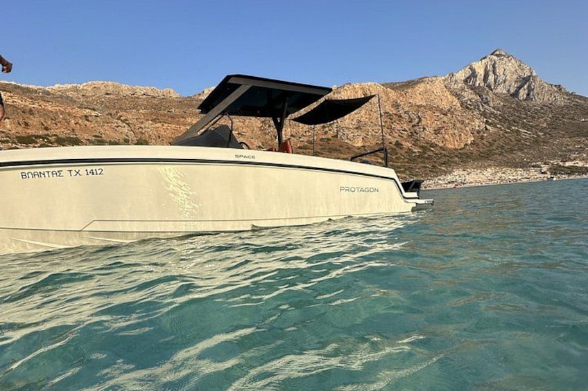 Ein Boot mit dem Namen 'BNANTRE TX 1412', auf dessen Seite 'PROTAGON' steht, treibt auf klarem, blauem Wasser, im Hintergrund ein felsiger Berg. 2