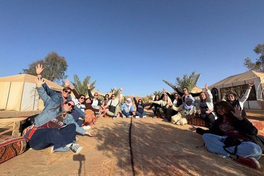 A group of people sitting on the ground in an outdoor setting with tents and palm trees in the background. 4