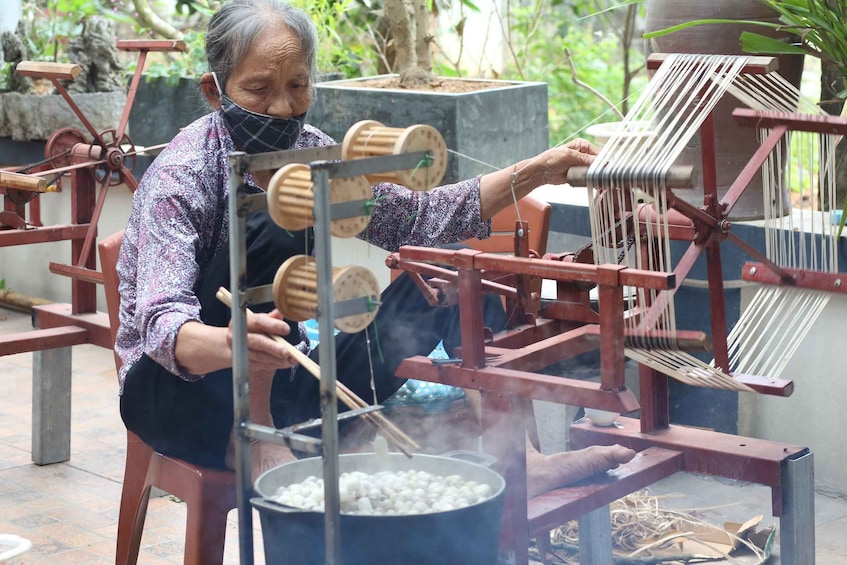 An individual operating a traditional spinning wheel, with a pot of boiling water and a basket of white items nearby. 1