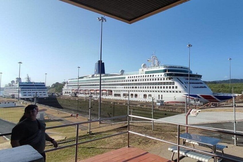 A large cruise ship docked at a port with a clear sky and a person sitting on a bench. 3