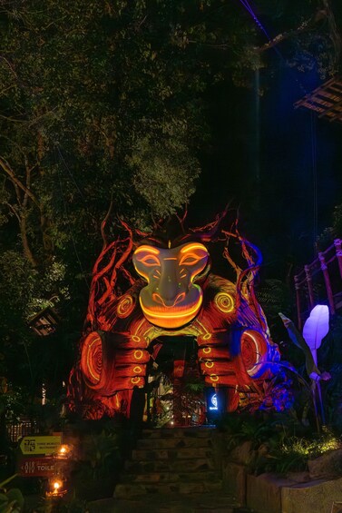 A large illuminated sculpture resembling a face with a mask, surrounded by trees and a wooden structure. 2
