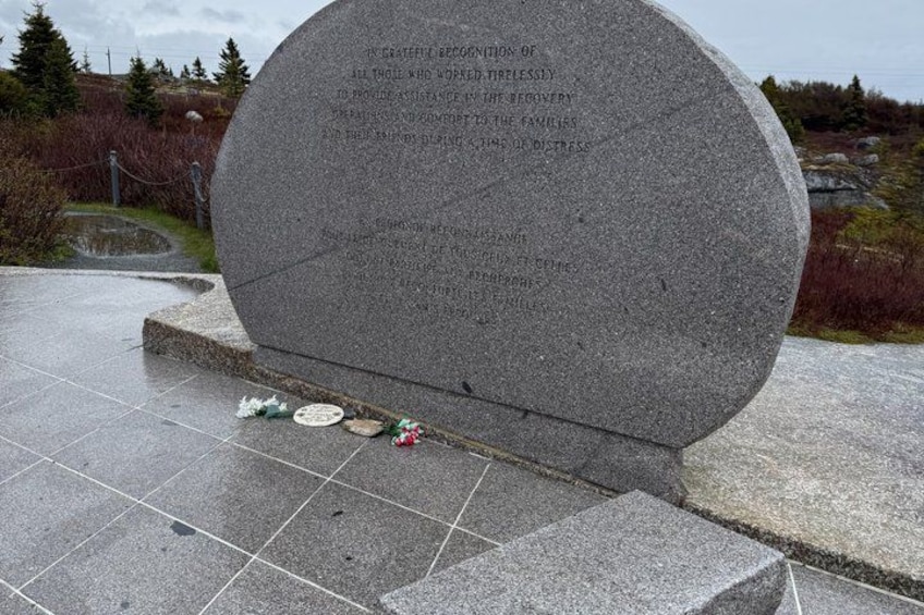A granite memorial with engraved text, surrounded by a paved area and some greenery. 3