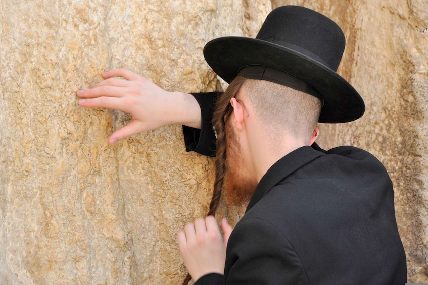 A person wearing a black hat and suit, touching a textured stone wall. 3
