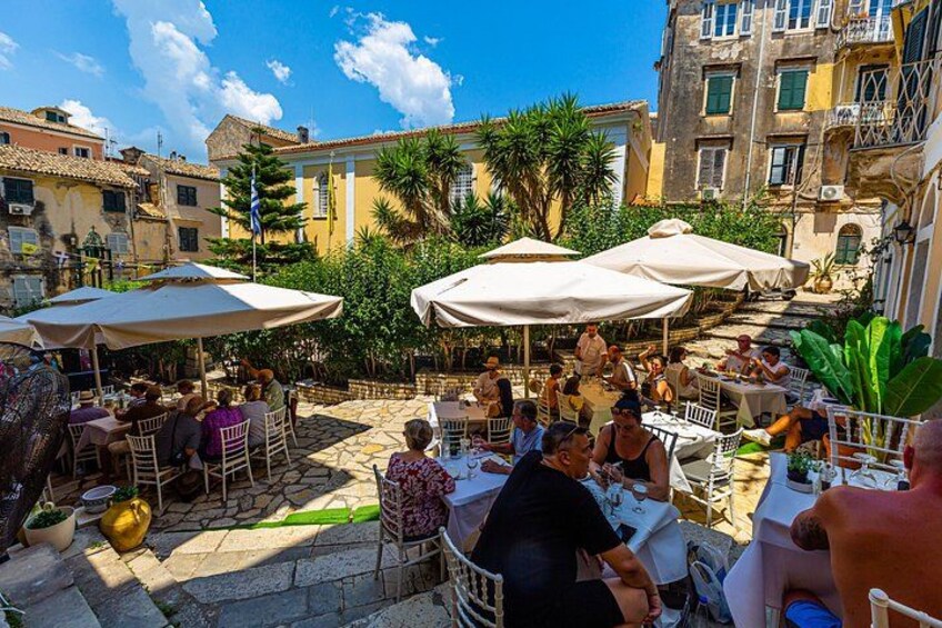 Un café al aire libre con mesas y sombrillas, rodeado de edificios y follaje. 3