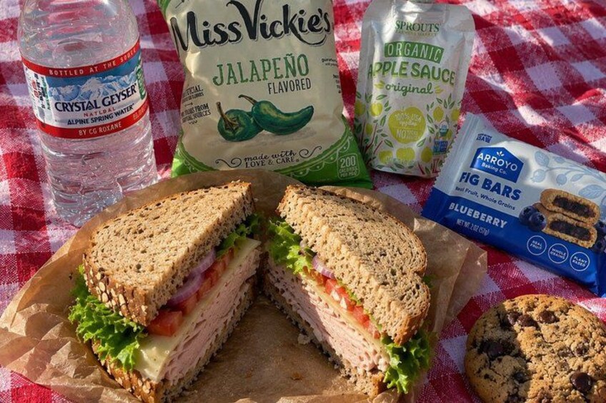 A sandwich cut in half, a bottle of water, a bag of jalapeño flavored seasoning, a bag of organic apple sauce, and a package of blueberry fig bars. 3