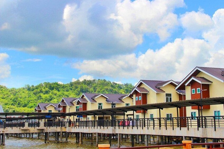 A row of yellow buildings with red roofs on a wooden pier over water, with a green hill in the background. 3