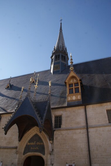 A historic building with a pointed roof, a tower with a cross, and a sign reading 'hostel-vieu 1443.' 1