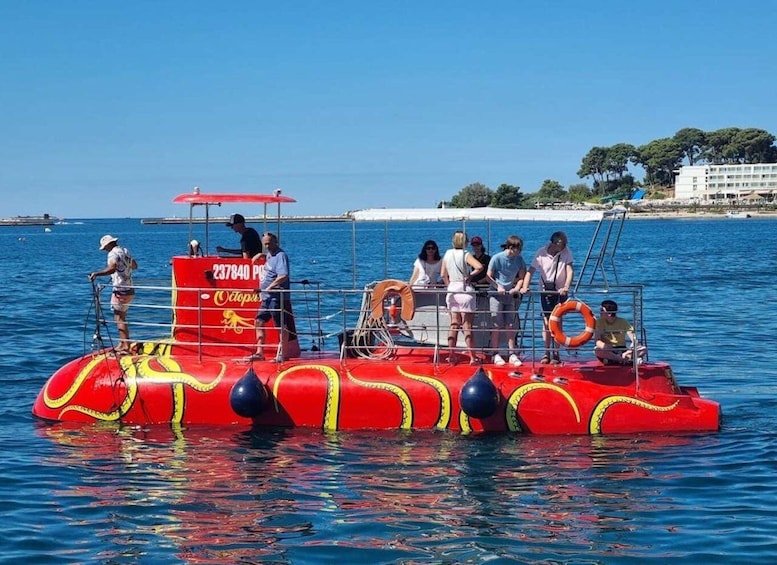 A red boat with passengers and a lifebuoy, floating on calm water with a clear sky and distant buildings. 1