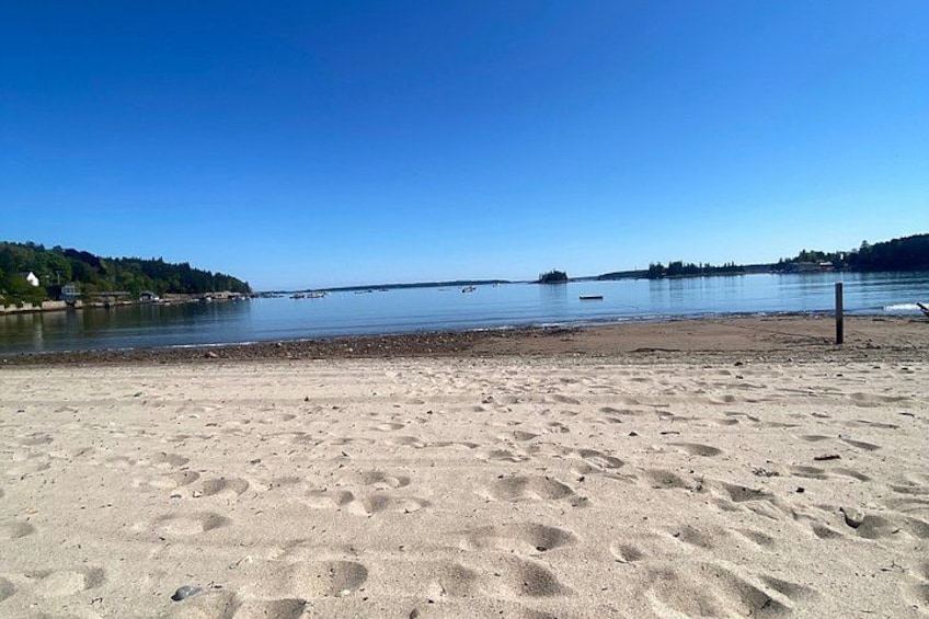 Une plage de sable avec des empreintes, une eau calme et une côte lointaine bordée d’arbres. 3