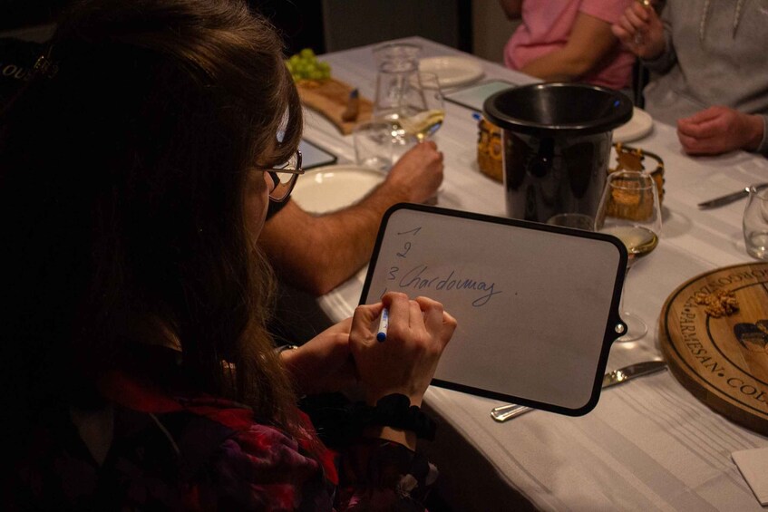 A person writing on a clipboard with a list, a wine glass, and a bottle of wine on a table. 4