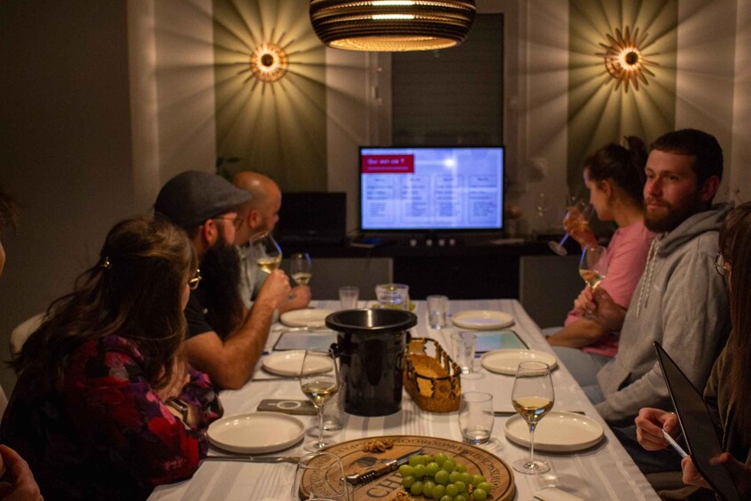 A group of people dining at a table with a laptop, wine glasses, and a basket of grapes. 5
