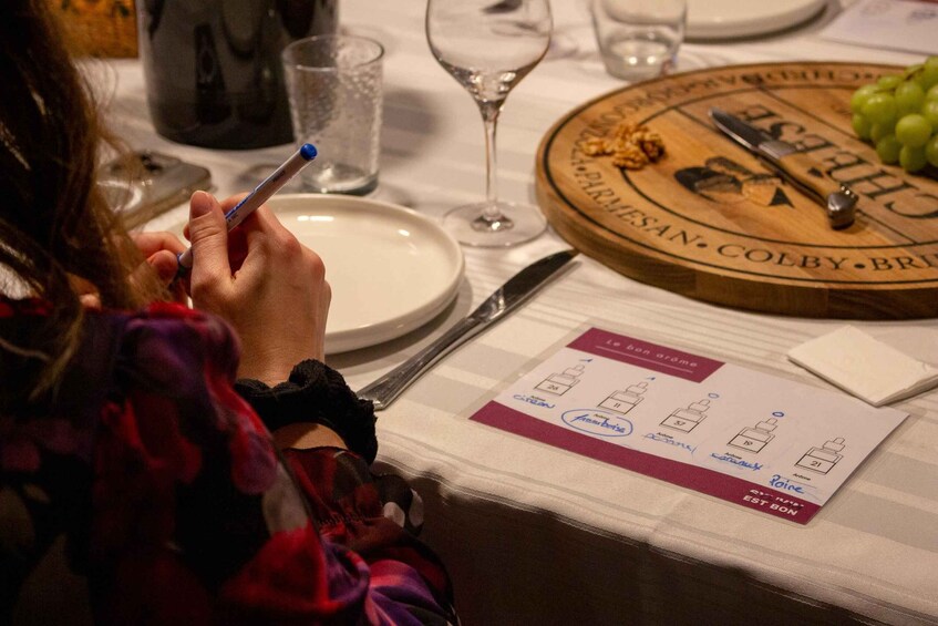 A person writing on a menu at a restaurant table with a wine glass and a wooden board with cheese and grapes. 2