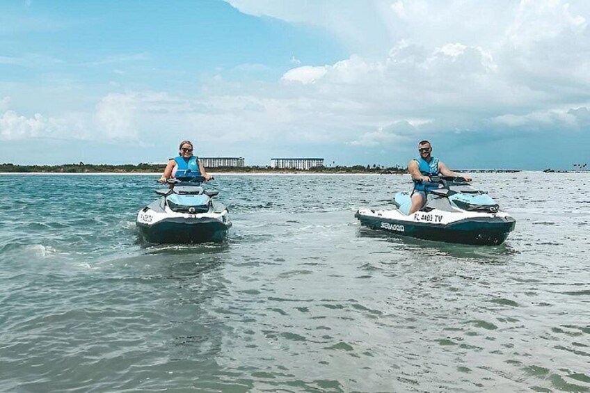 Two people riding jet skis on calm water with a pier and buildings in the background. 4
