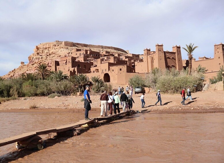 A group of people crossing a wooden bridge over a river, with traditional buildings and palm trees in the background. 2
