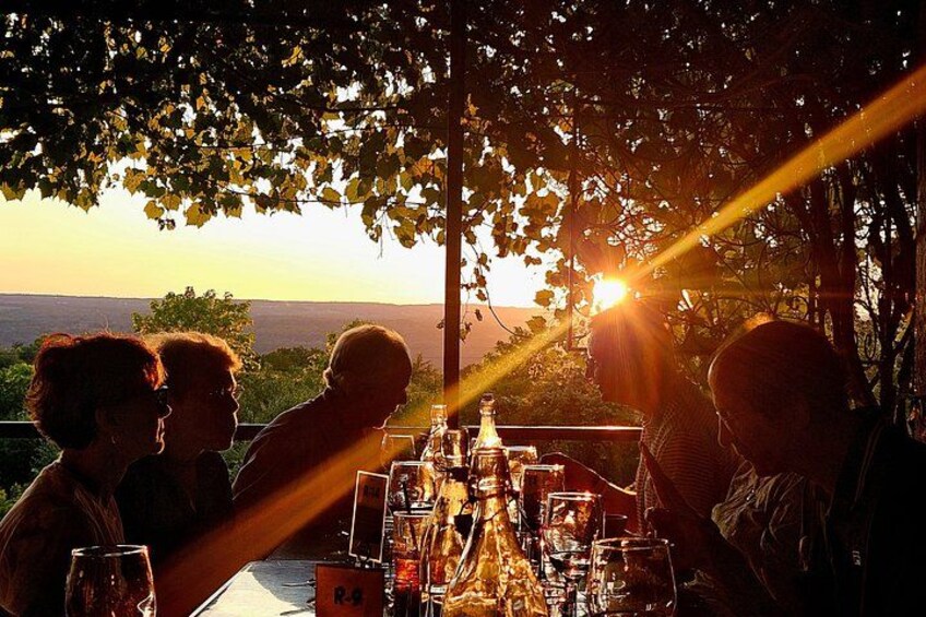 A group of people dining outdoors at sunset with a table set for a meal, surrounded by trees and a view of the landscape. 2