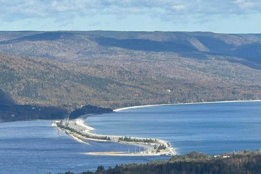 A coastal landscape with a curved shoreline, a breakwater, and forested hills in the background. 4