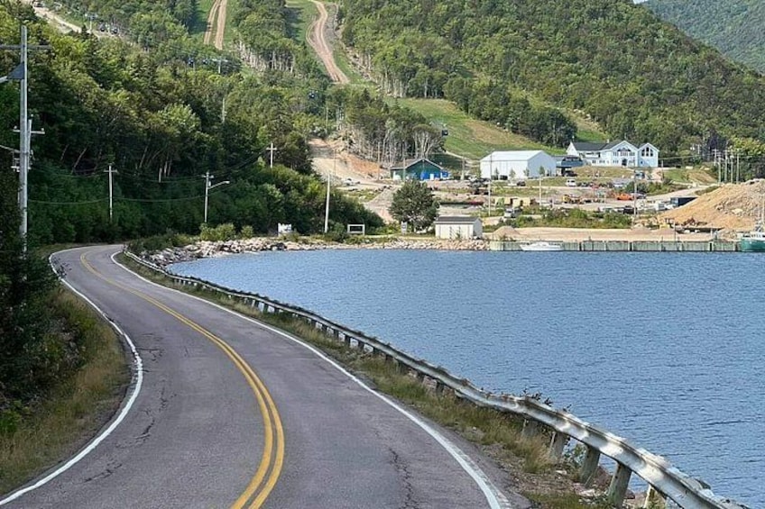 A winding road curving alongside a body of water, with houses and a hill in the background. 2