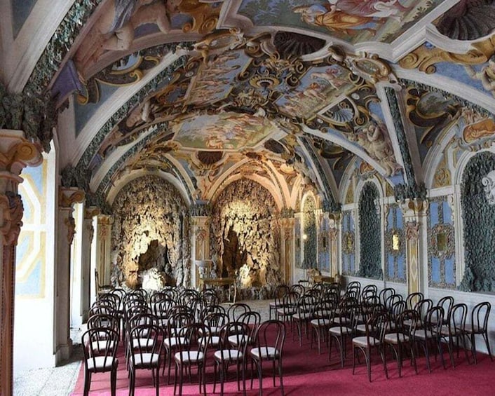 An ornate hall with a high ceiling, intricate frescoes, and rows of chairs. 1