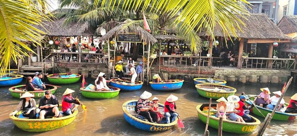A group of people in traditional boats on a river, with a restaurant and palm trees in the background. 5