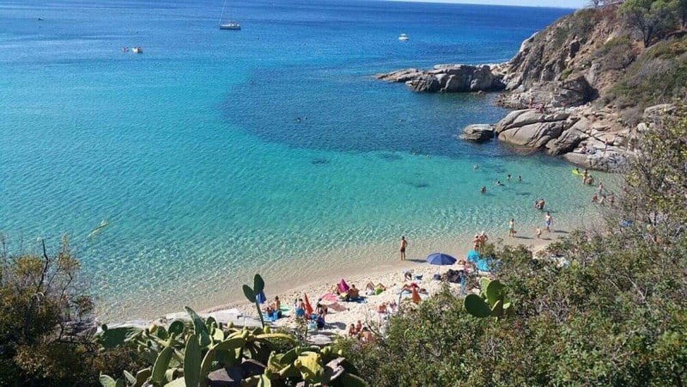A coastal beach with clear turquoise water, rocky outcrops, and people sunbathing and swimming. 5