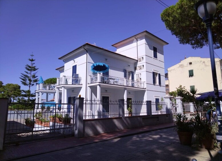 A white two-story building with balconies, a blue awning, and a fence with potted plants. 2