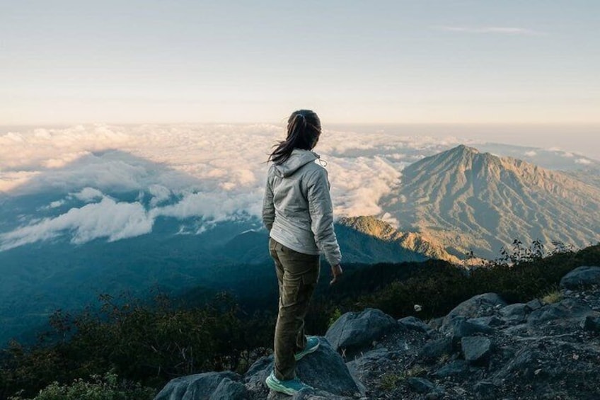 Une personne se tient debout sur un promontoire rocheux, d’où elle domine une chaîne de montagnes, avec des nuages en contrebas. 1