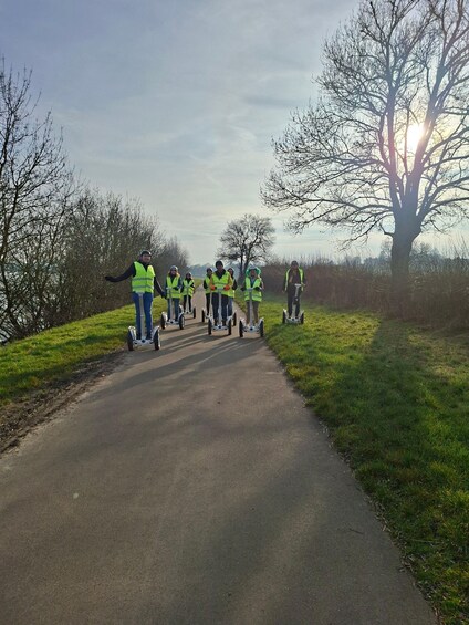 A group of people on electric scooters on a paved path surrounded by grass and trees. 1
