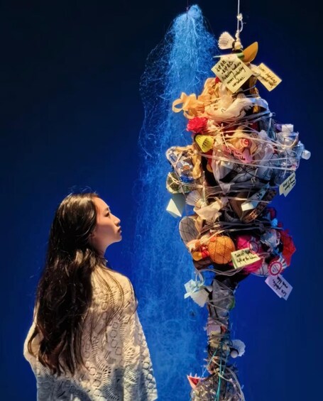 A person standing in front of a large, colorful, vertically hanging structure with various items attached, including tags and notes. 2